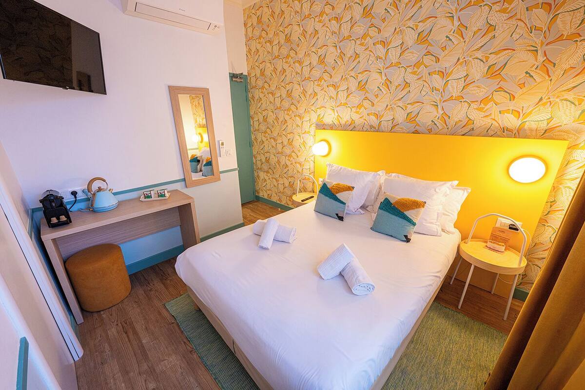 A hotel room with a bed, bedside table, lamp, and a desk with a kettle and a mirror.