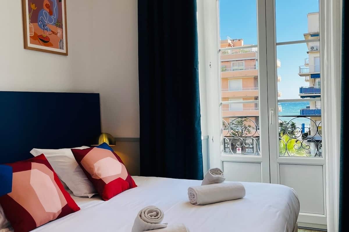 A hotel room with a large bed, a painting on the wall, and a view of buildings through the window.