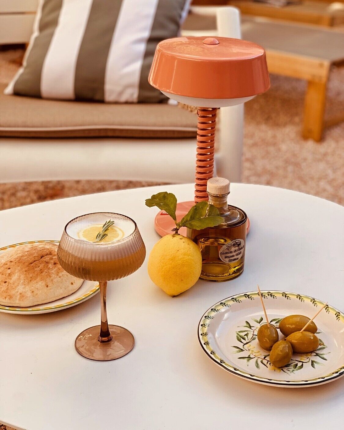 A table with a glass of a yellow beverage, a lemon, a bottle of olive oil, and a plate with olives.