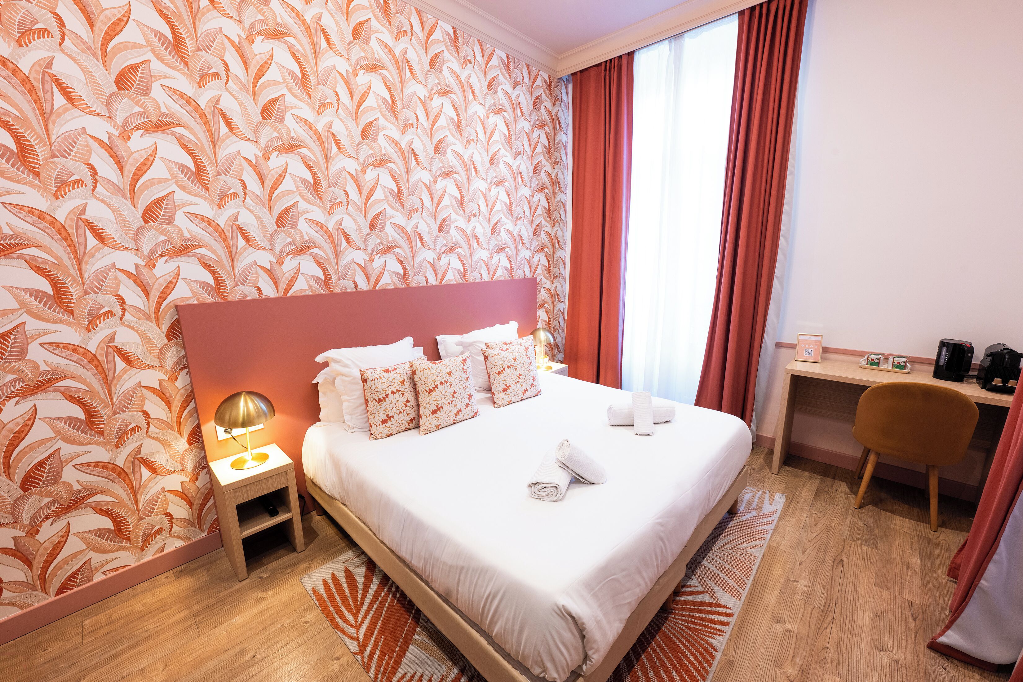 A hotel room with a large bed, a desk, a chair, and a nightstand with a lamp.