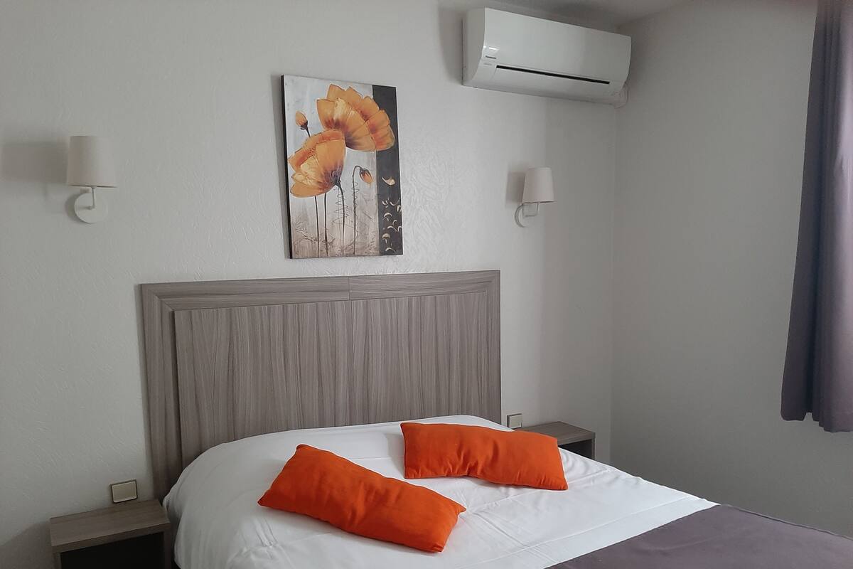 Habitación de hotel con una cama, dos almohadas naranjas, un cabecero de madera, una pared blanca y un equipo de aire acondicionado.