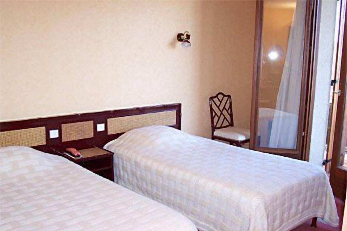 Dos camas individuales con cabeceros de madera, una mesita de noche pequeña, una silla y una ventana con cortina.