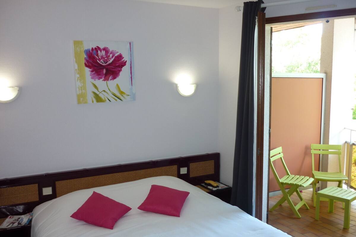 Habitación de hotel con cama, dos almohadas, mesita de noche, lámpara, un cuadro y balcón con sillas.