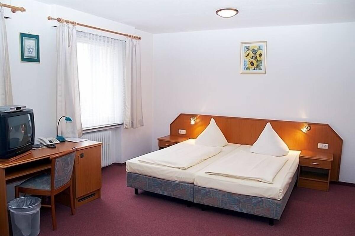 A hotel room with a double bed, a desk with a television, a chair, a trash bin, and a window with curtains.