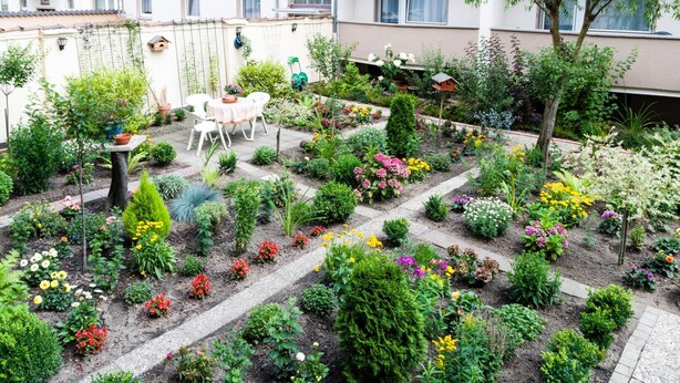 En velholdt have med forskellige planter og blomster, et lille siddeområde med bord og stole, samt en bygning i baggrunden.