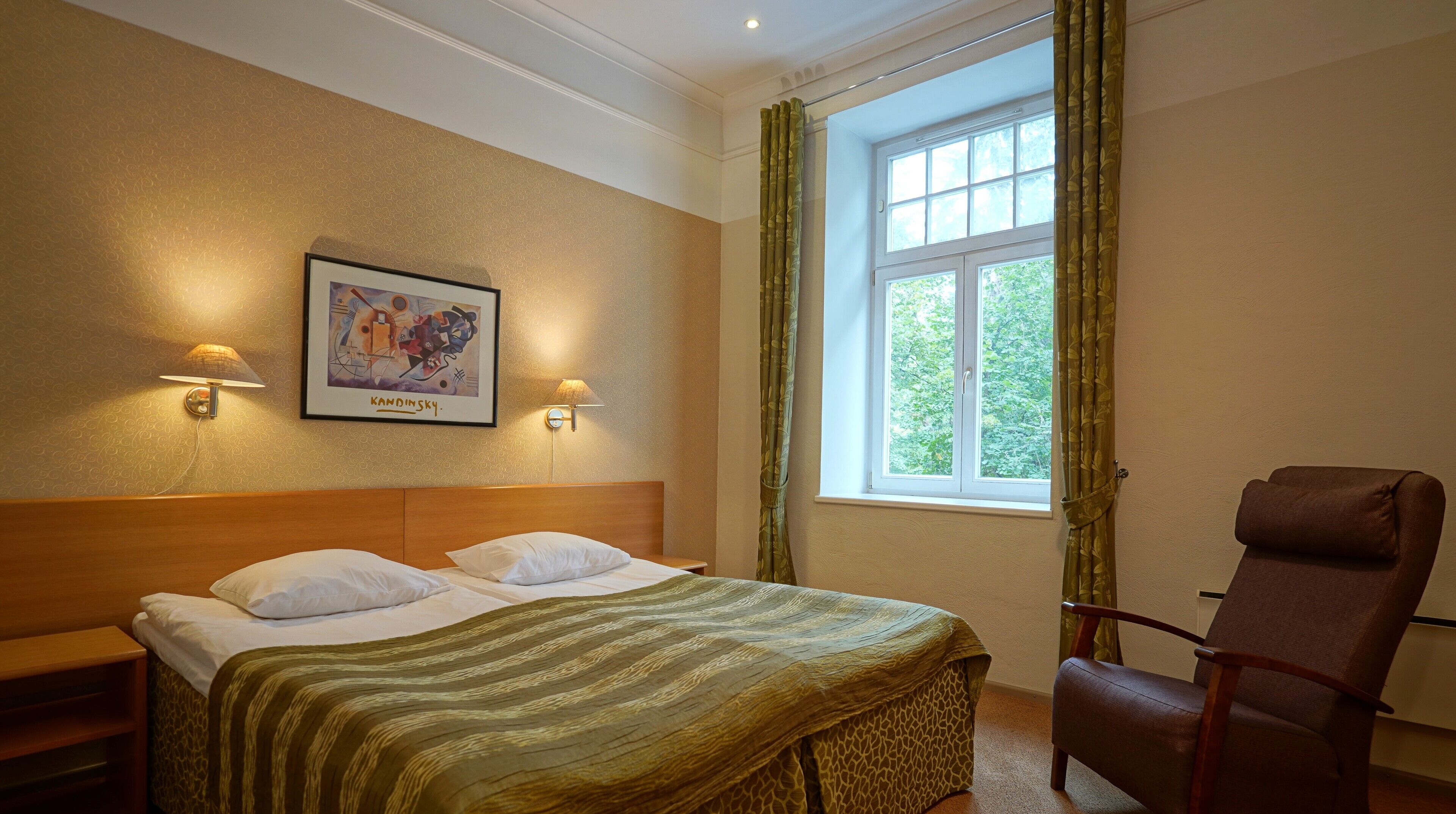Una habitación de hotel con una cama, dos lámparas de pared, una obra de arte enmarco, una ventana con cortinas y una silla.