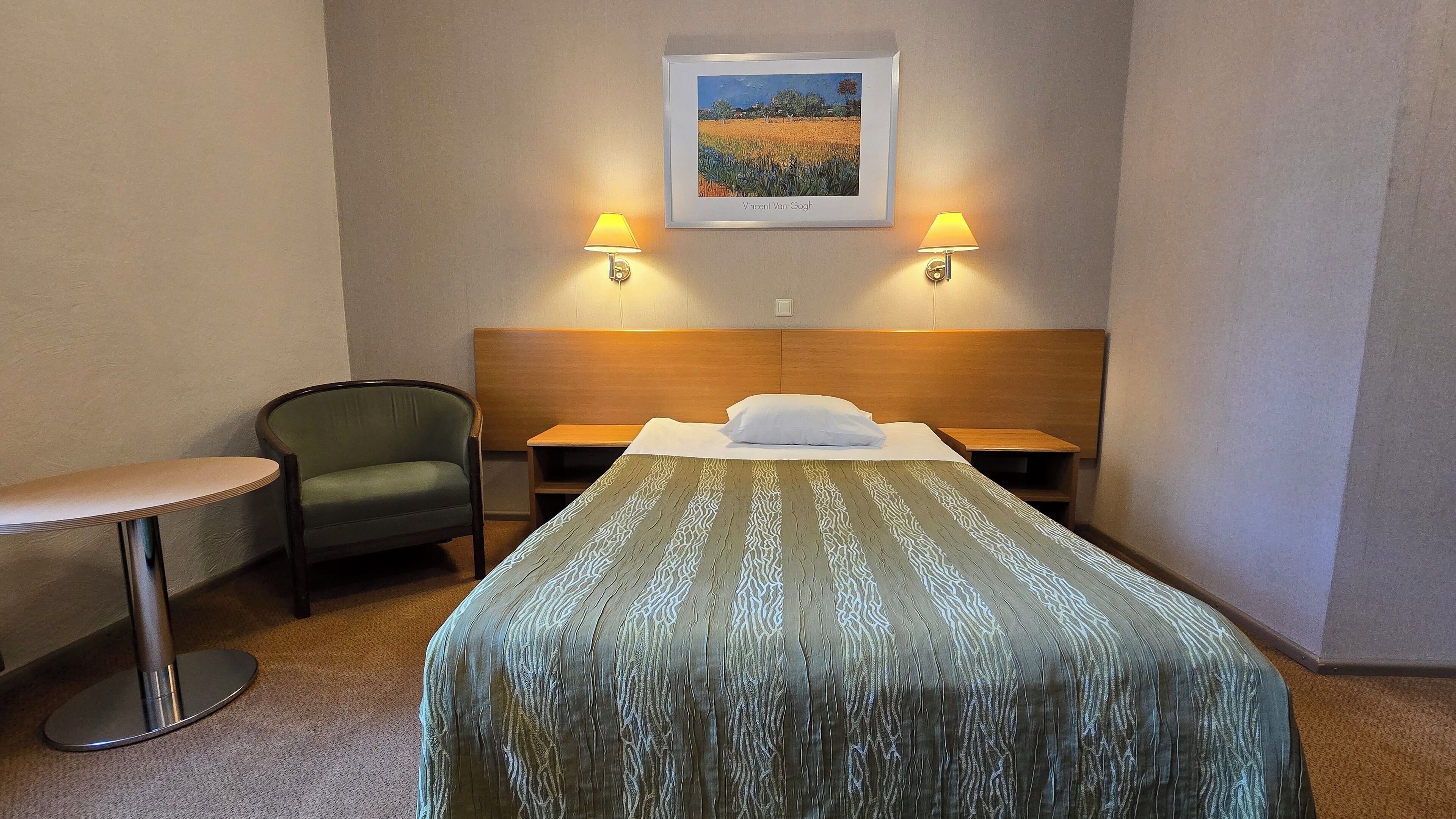 Habitació d'hotel amb un llit, una cadira, una taula petita, dues làmpades penjades a la paret i una fotografia amb marc.