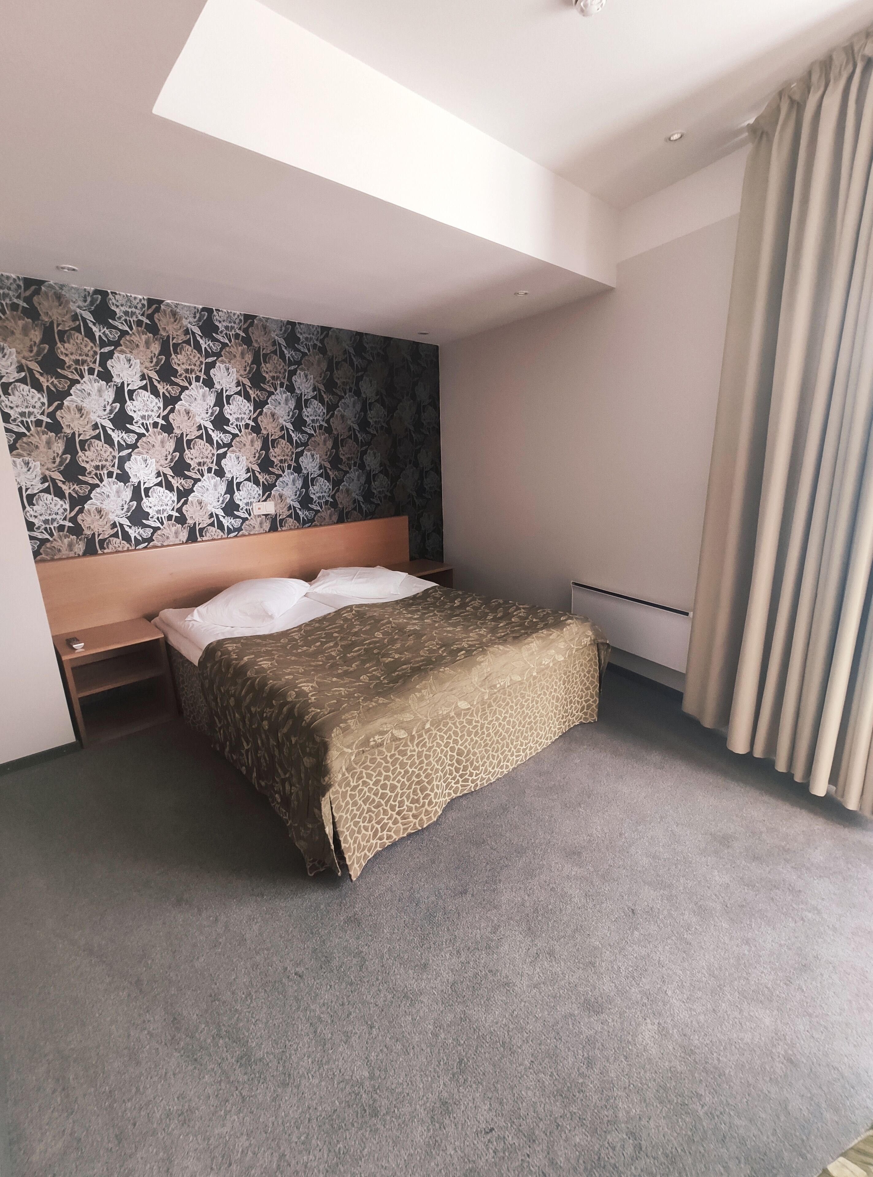 A hotel room with a bed, a headboard, a nightstand, a wall with a floral pattern, and a window with curtains.