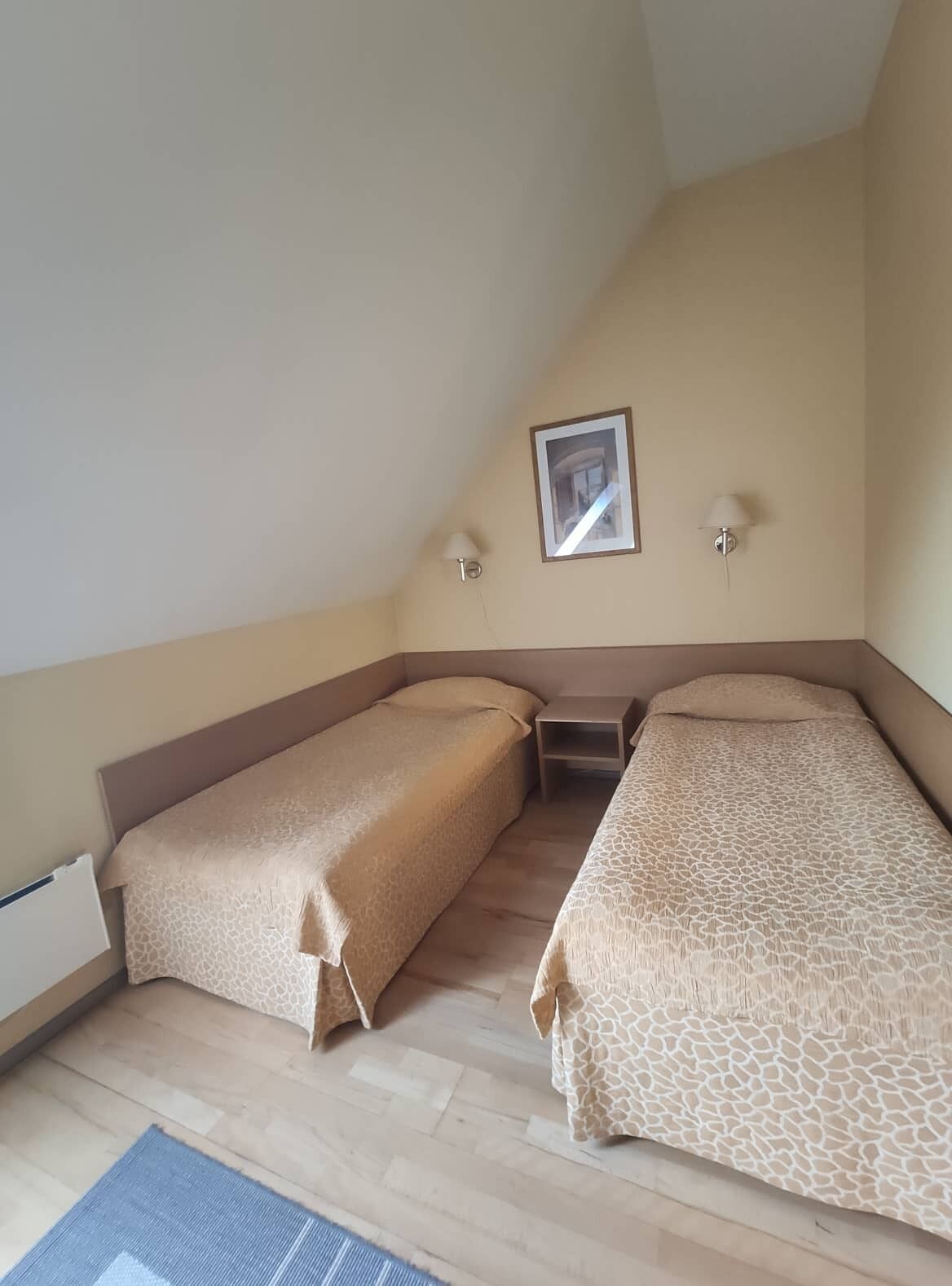 A small, single-room with two beds, a small table, and a picture on the wall.