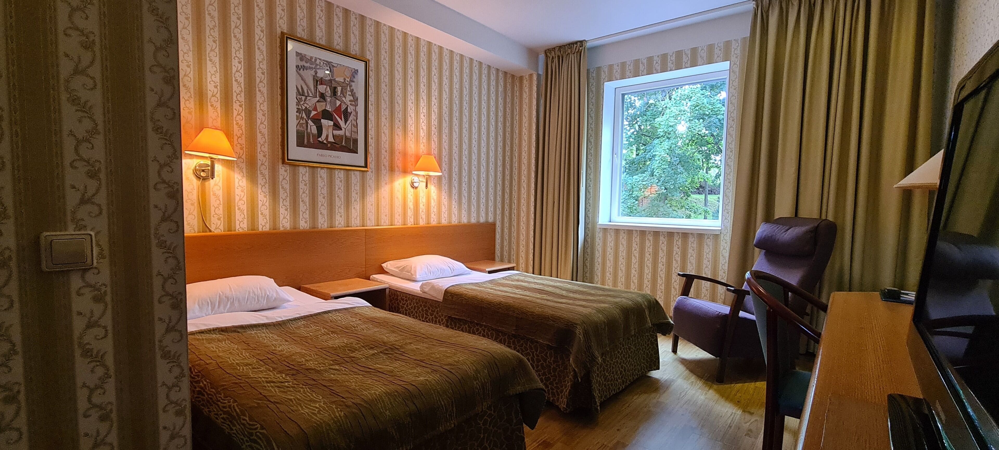 Quarto de hotel com duas camas, uma cabeceira de madeira, televisão, uma cadeira e janela com cortinas.