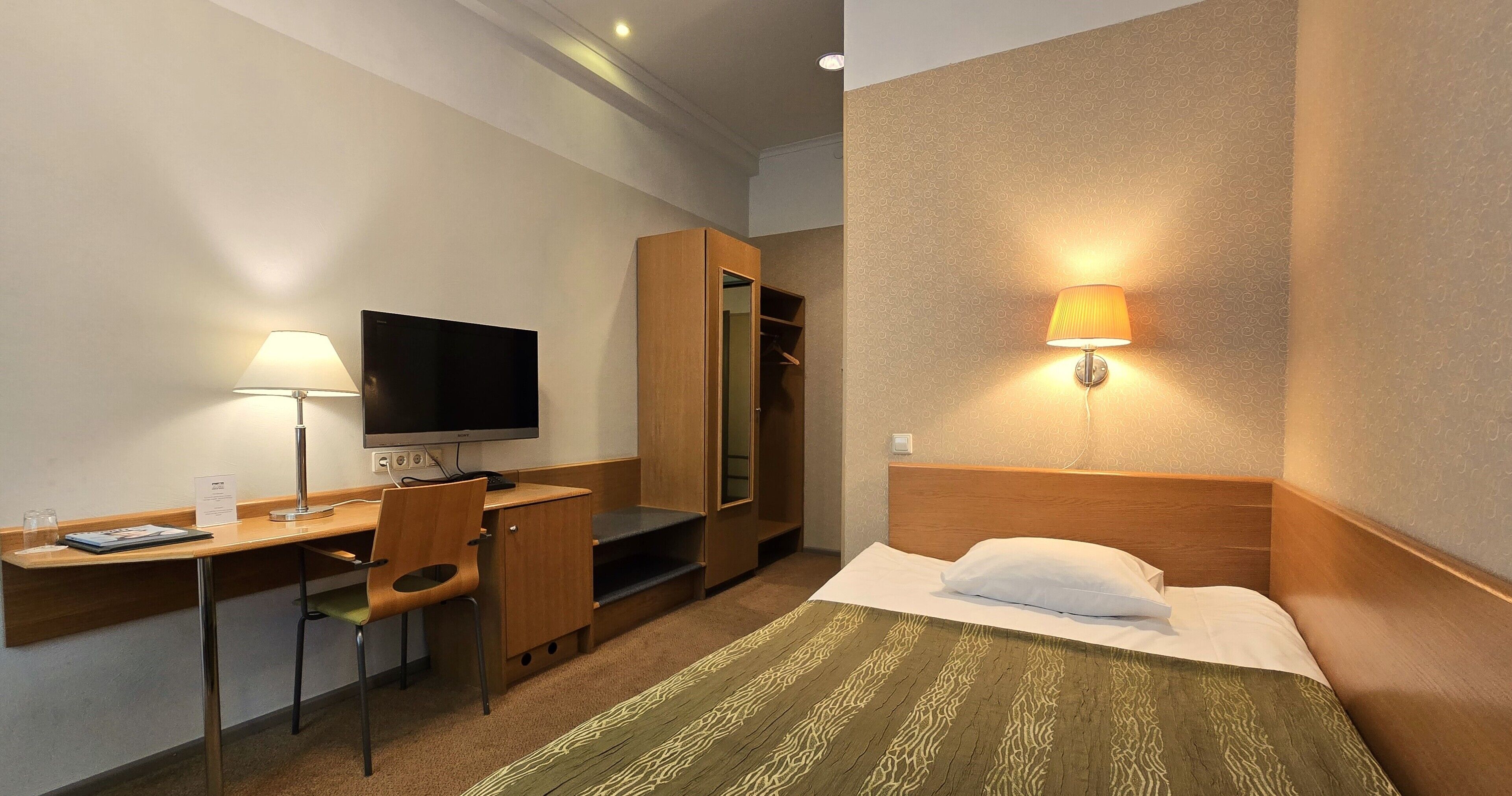 Une chambre d’hôtel comprenant un lit, un bureau avec une chaise, une télévision, une armoire et une lampe fixée au mur.
