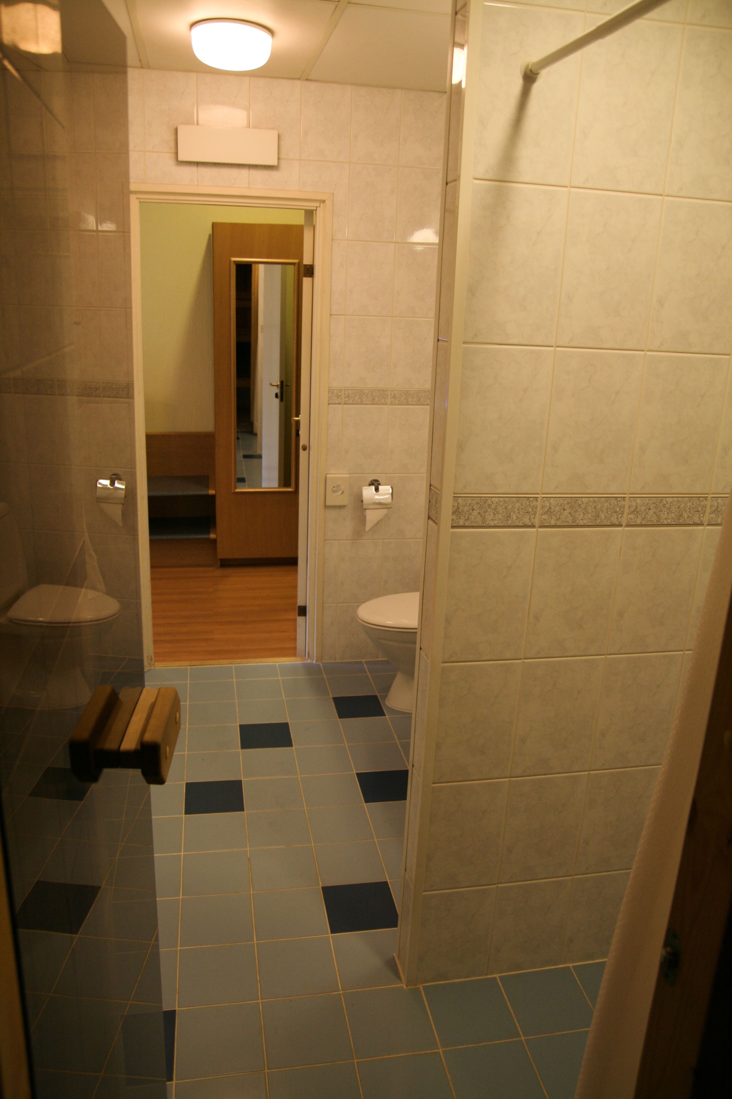 Un baño con inodoro, lavabo y cabina de ducha. Hay un banco de madera en la esquina. El suelo tiene un estampado de cuadros.