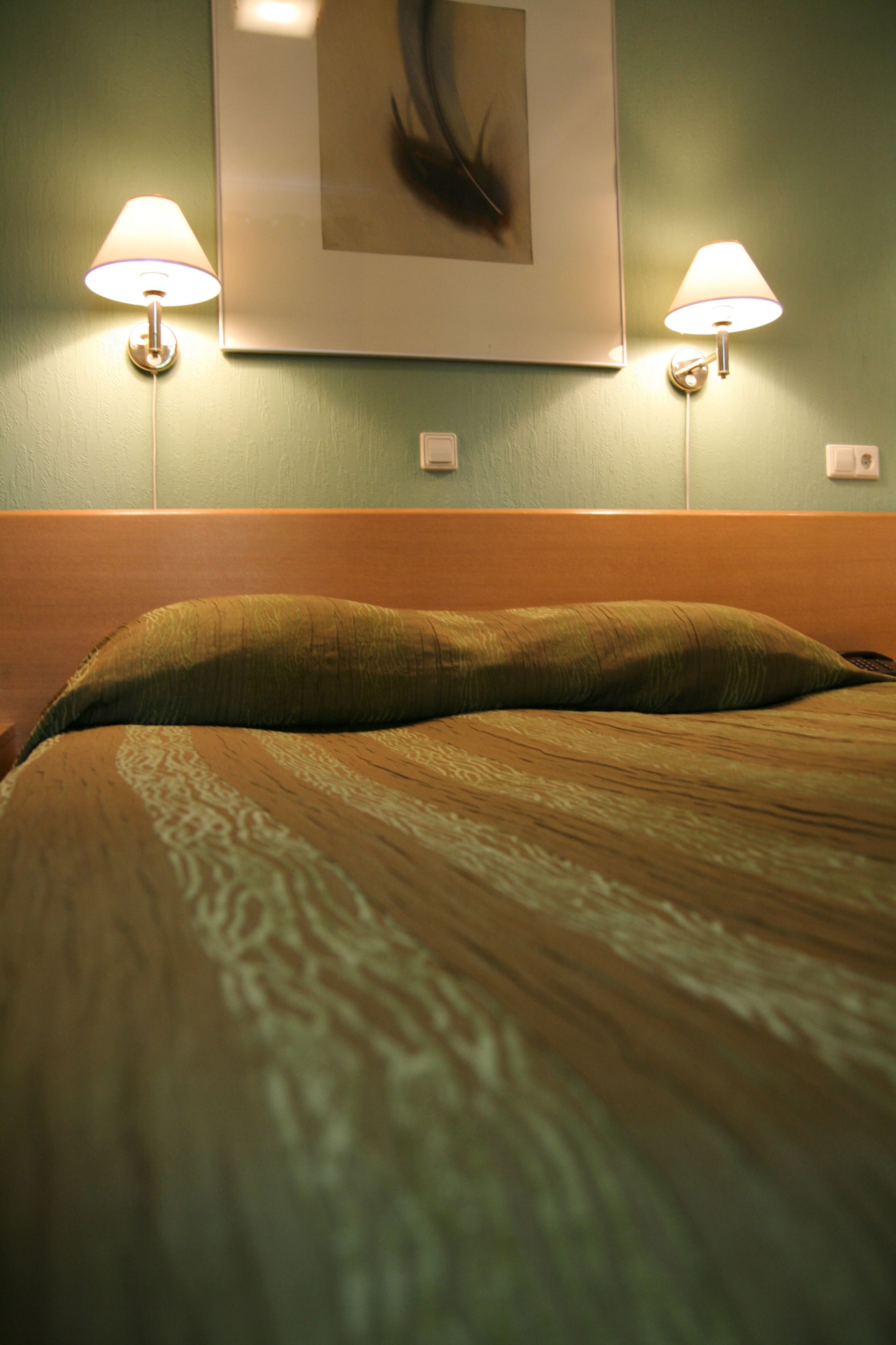 Una cama con cabecera, dos lámparas fijadas a la pared y una obra enmarcada sobre la cama.