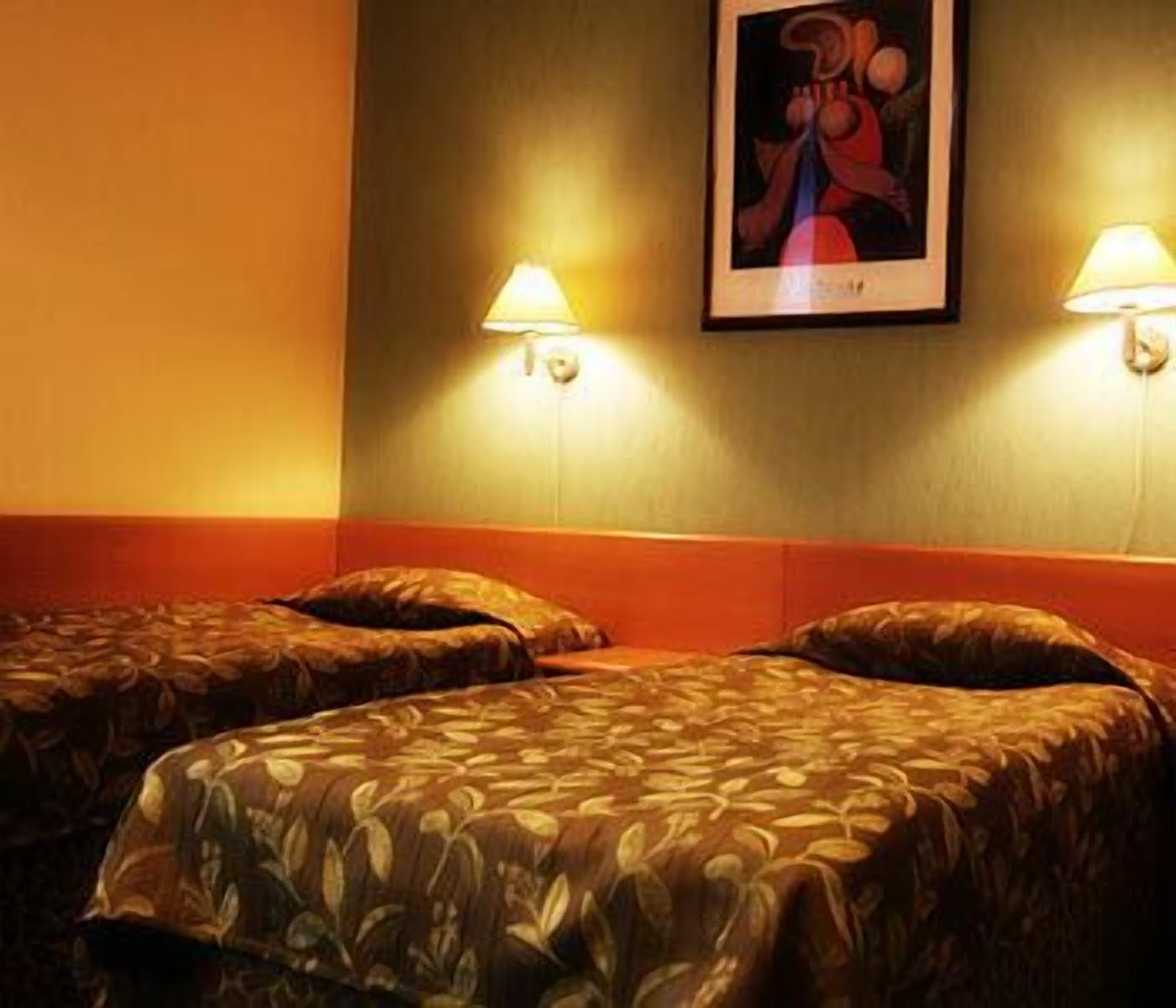 Hotellihuoneessa on kaksi sänkyä, joissa on molemmissa kuviollinen päiväpeite ja tyynyjä. Sänkyjen molemmilla puolilla on seinään kiinnitetyt lamput.