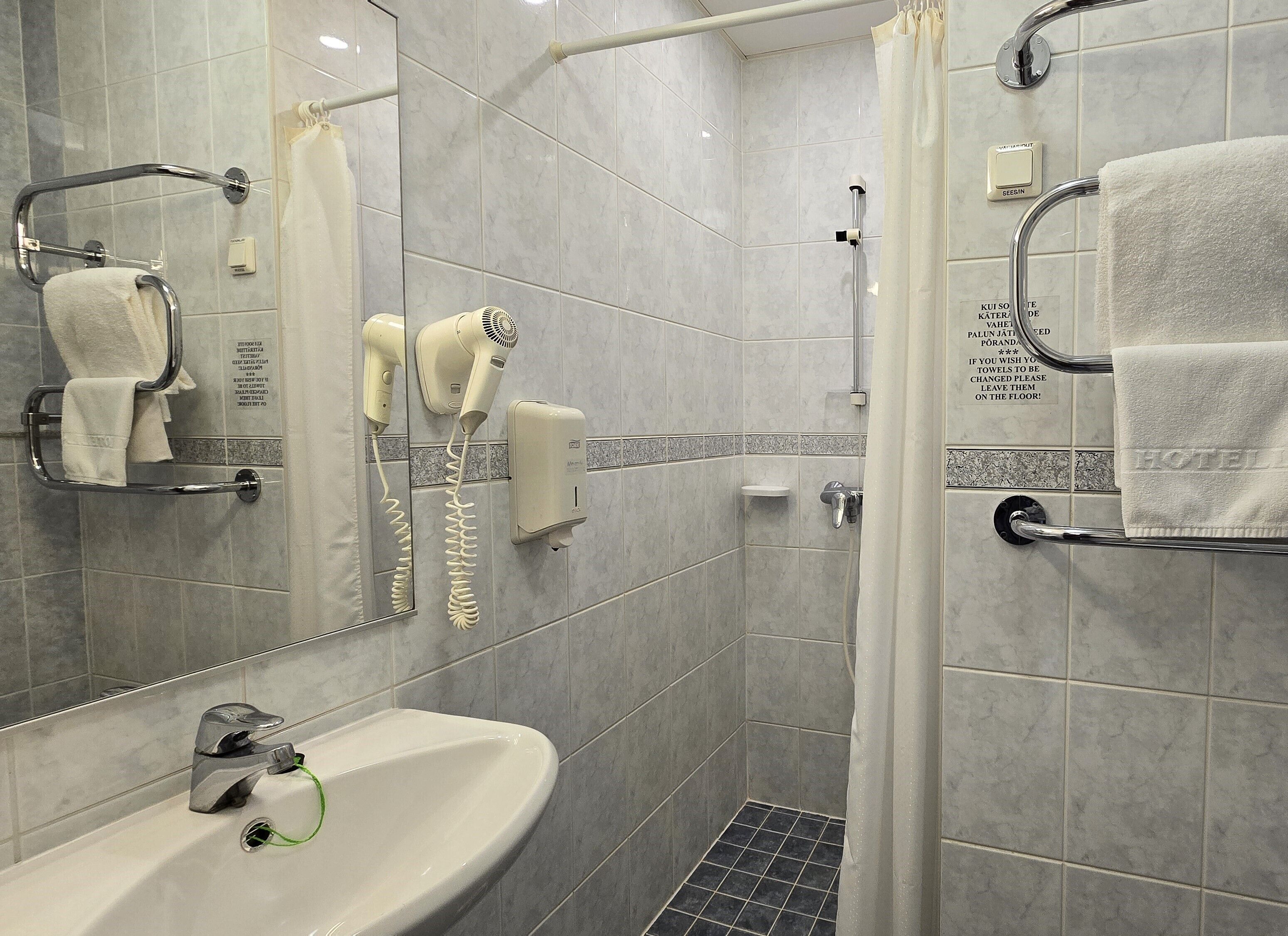 Une salle de bain d’hôtel équipée d’un lavabo, d’un miroir, d’un porte-serviettes, d’un sèche-cheveux et d’un rideau de douche.
