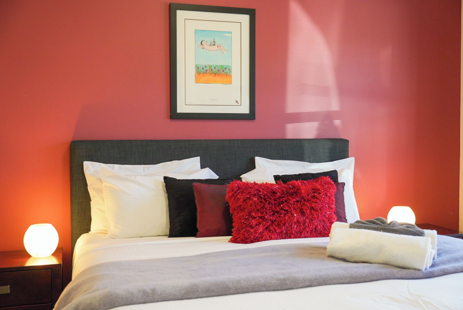 Uma cama bem arrumada com almofadas brancas e vermelhas, um cobertor cinza e um quadro na parede.