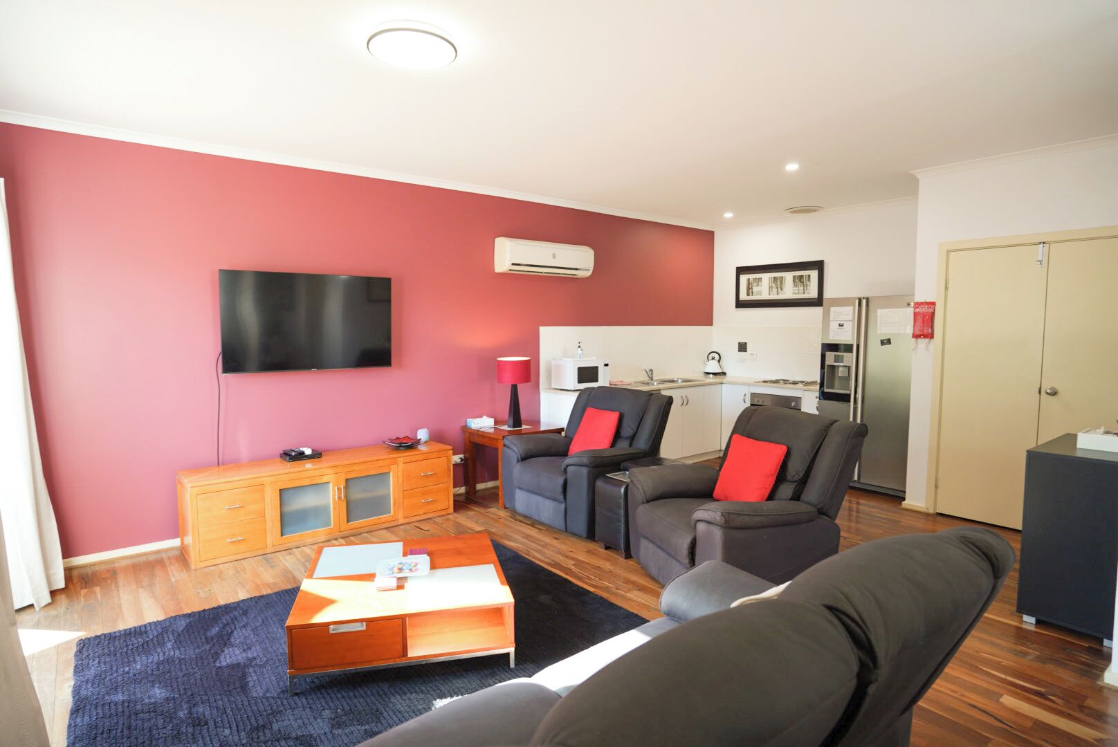 A modern living room with a flat-screen TV, a sofa set, a wooden coffee table, and a kitchen area in the background.