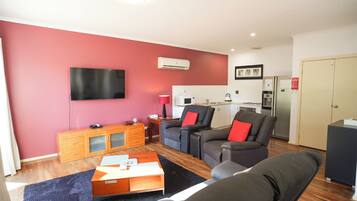 A modern living room with a flat-screen TV, a sofa set, a wooden coffee table, and a kitchen area in the background.