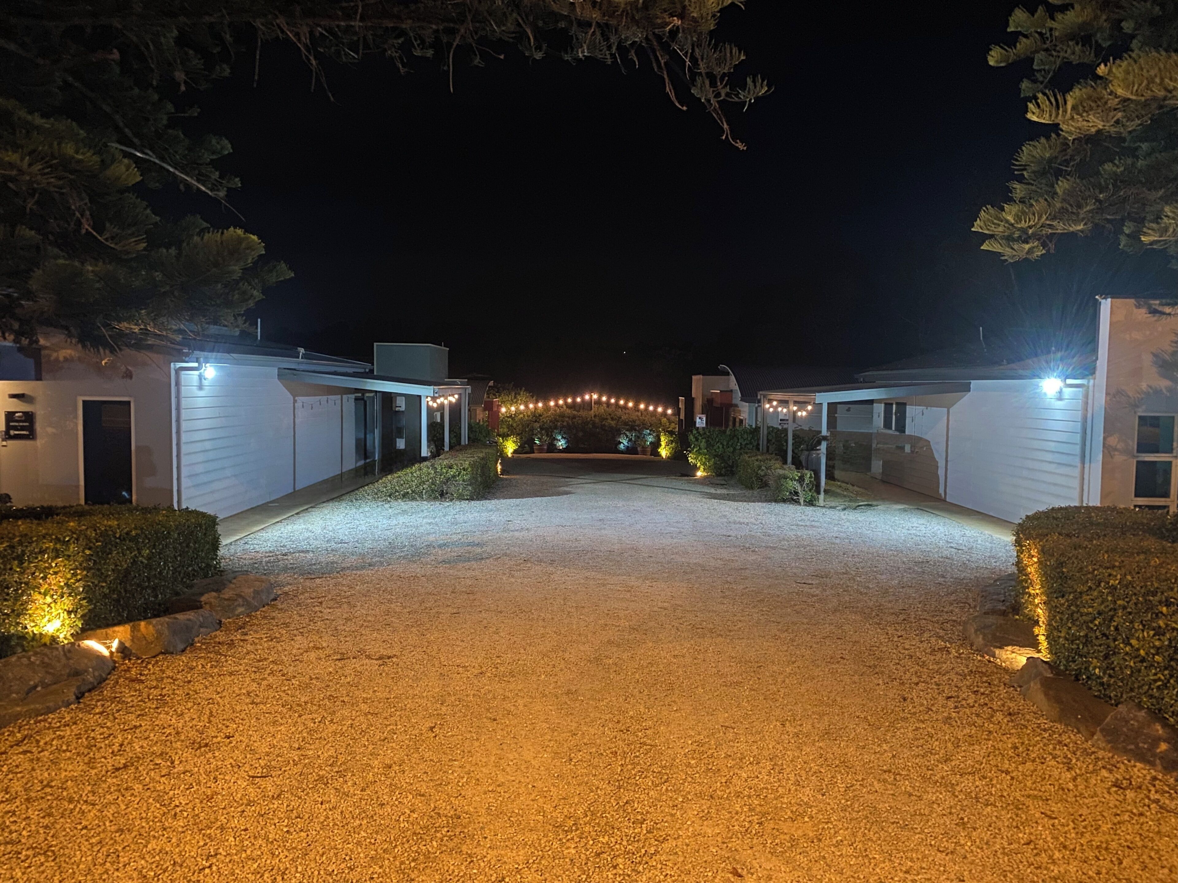 A gravel driveway leading to a building with a covered entrance, surrounded by hedges and illuminated by string lights.