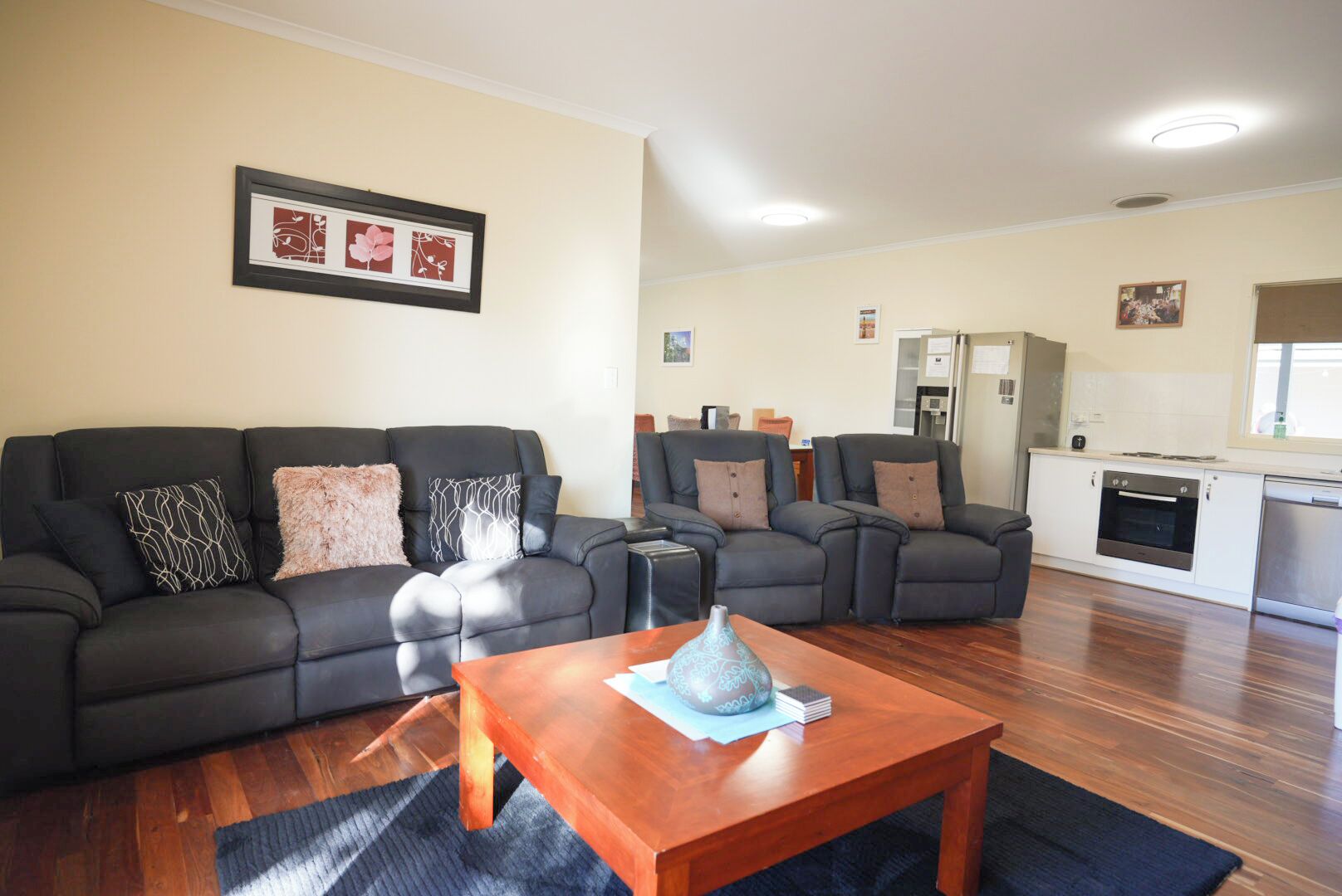 A living room with a black sofa set, a wooden coffee table, and a kitchen area with white cabinets and appliances.