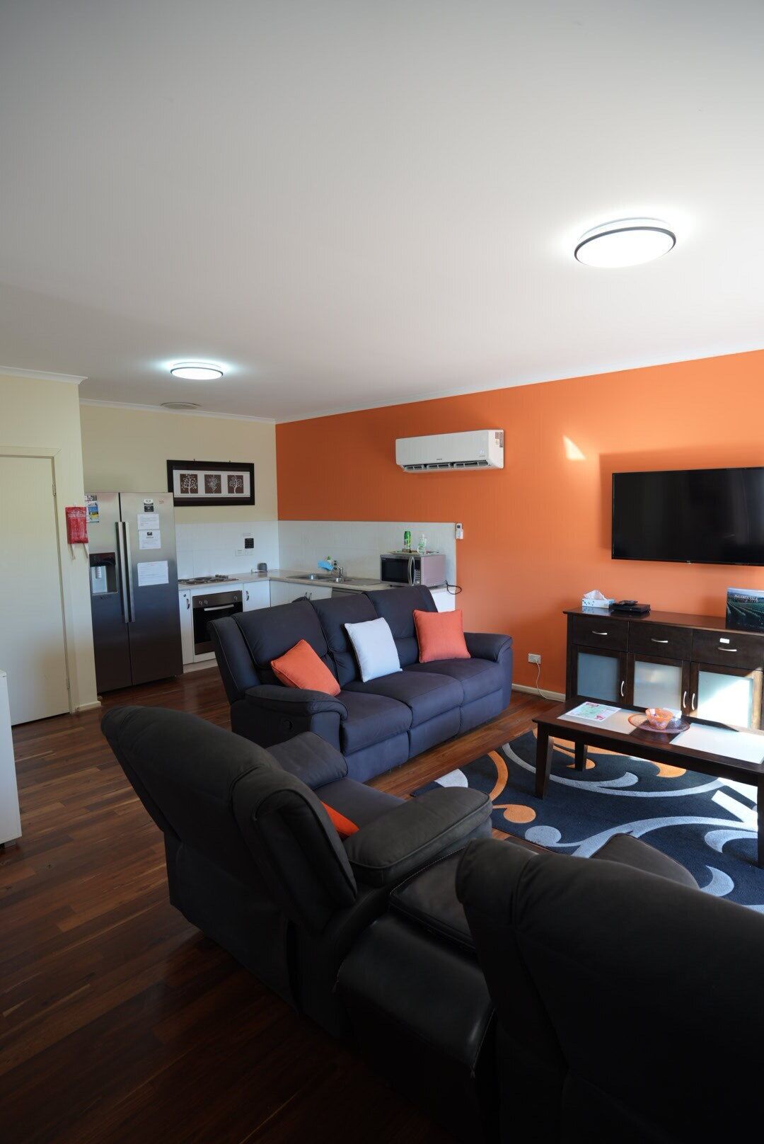 A modern living room with a dark blue sofa, a black leather chair, and a wooden coffee table. A kitchen with a refrigerator and stove is visible in the background.