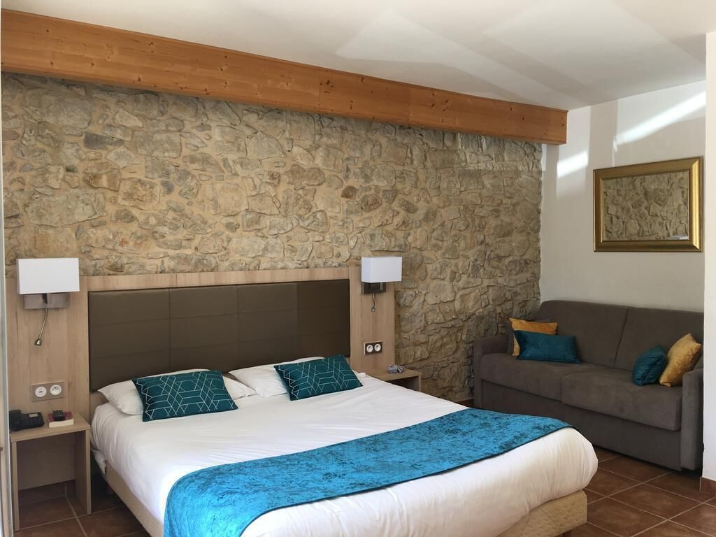 A bedroom with a stone wall, a bed with white bedding and blue accents, a wooden headboard, a nightstand with a lamp, and a gray sofa with cushions.