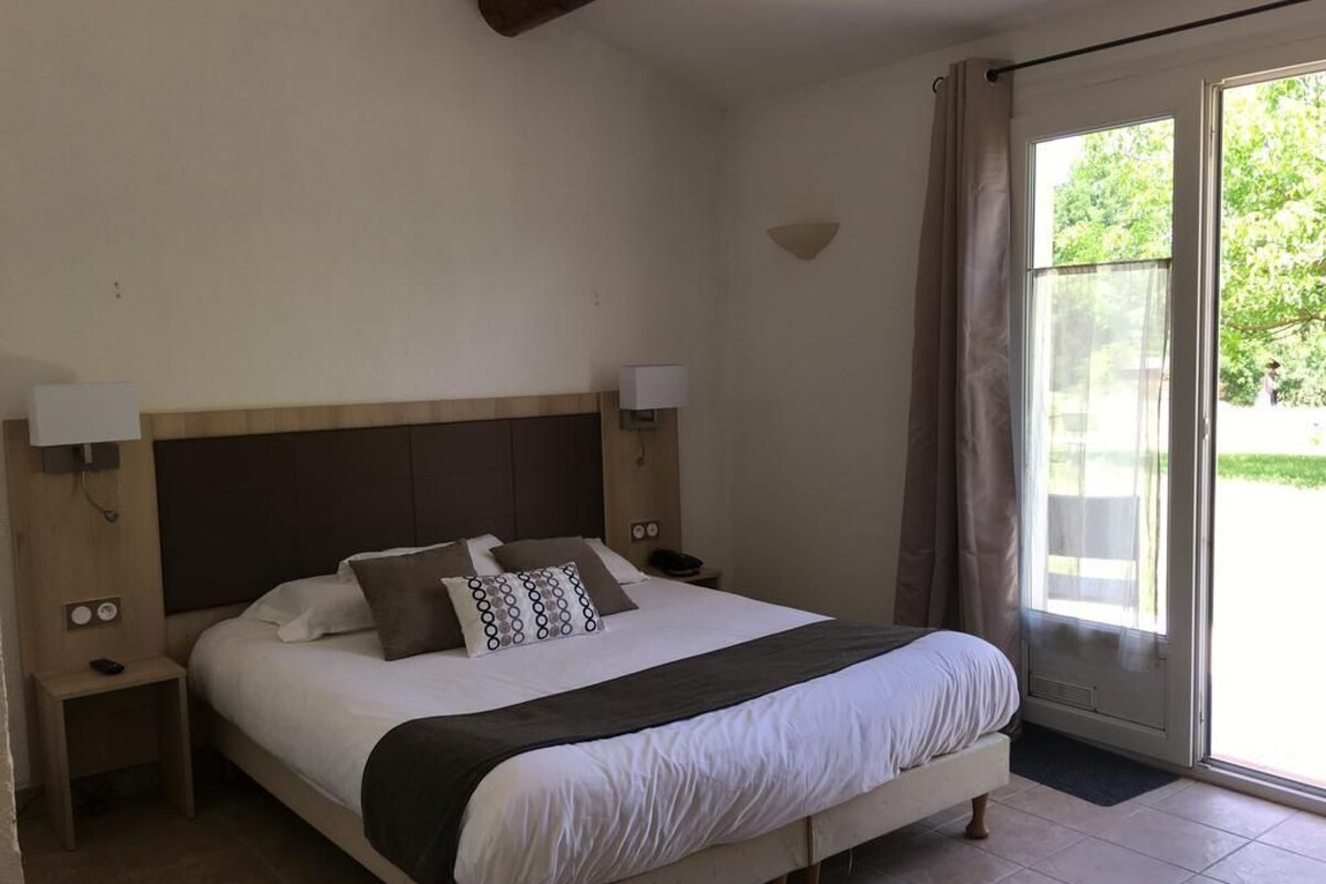 Quarto com cama, mesas de cabeceira, abajur, janela com cortinas e vista para o verde lá fora.