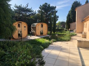 Two wooden barrel-shaped cabins with glass doors, surrounded by greenery and a paved walkway. - Déjeunez Sous l'Arbre (Rousset)