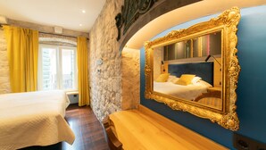 Une chambre d’hôtel avec un mur en pierre, un grand miroir décoratif, un banc en bois et un lit recouvert d’une courtepointe blanche.