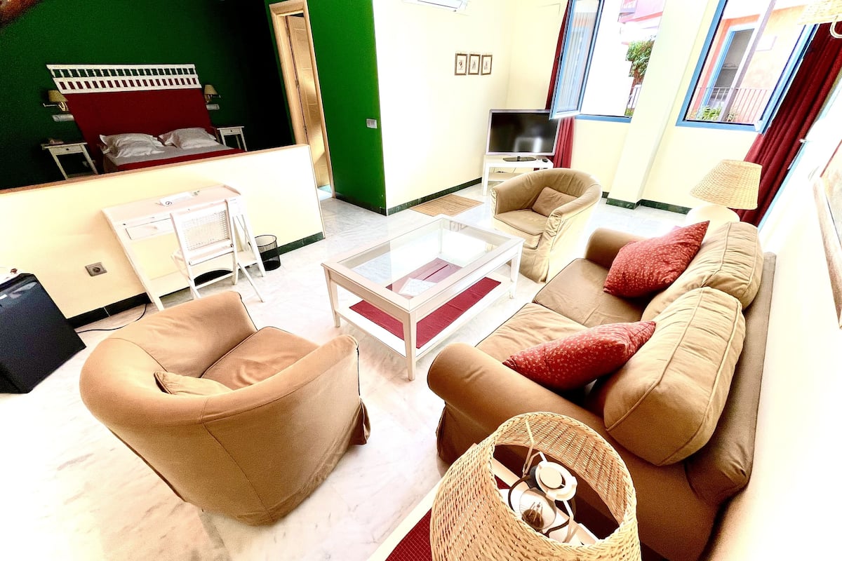 Sala de estar com sofá, poltrona e mesa de centro. Há uma televisão em um móvel e uma porta que dá para outro cômodo.