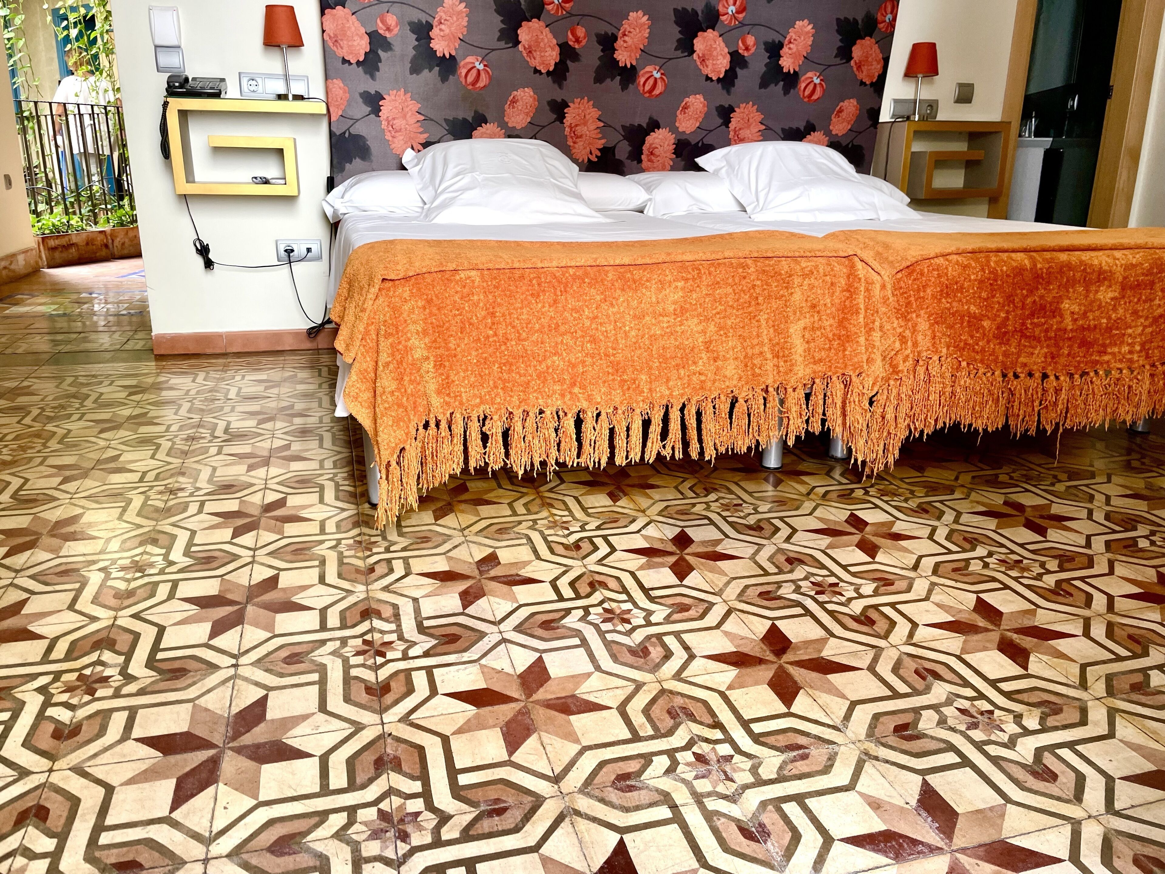 Une chambre d’hôtel avec un sol à motifs, un lit recouvert d’une couverture orange et une tête de lit recouverte d’un papier peint à fleurs.