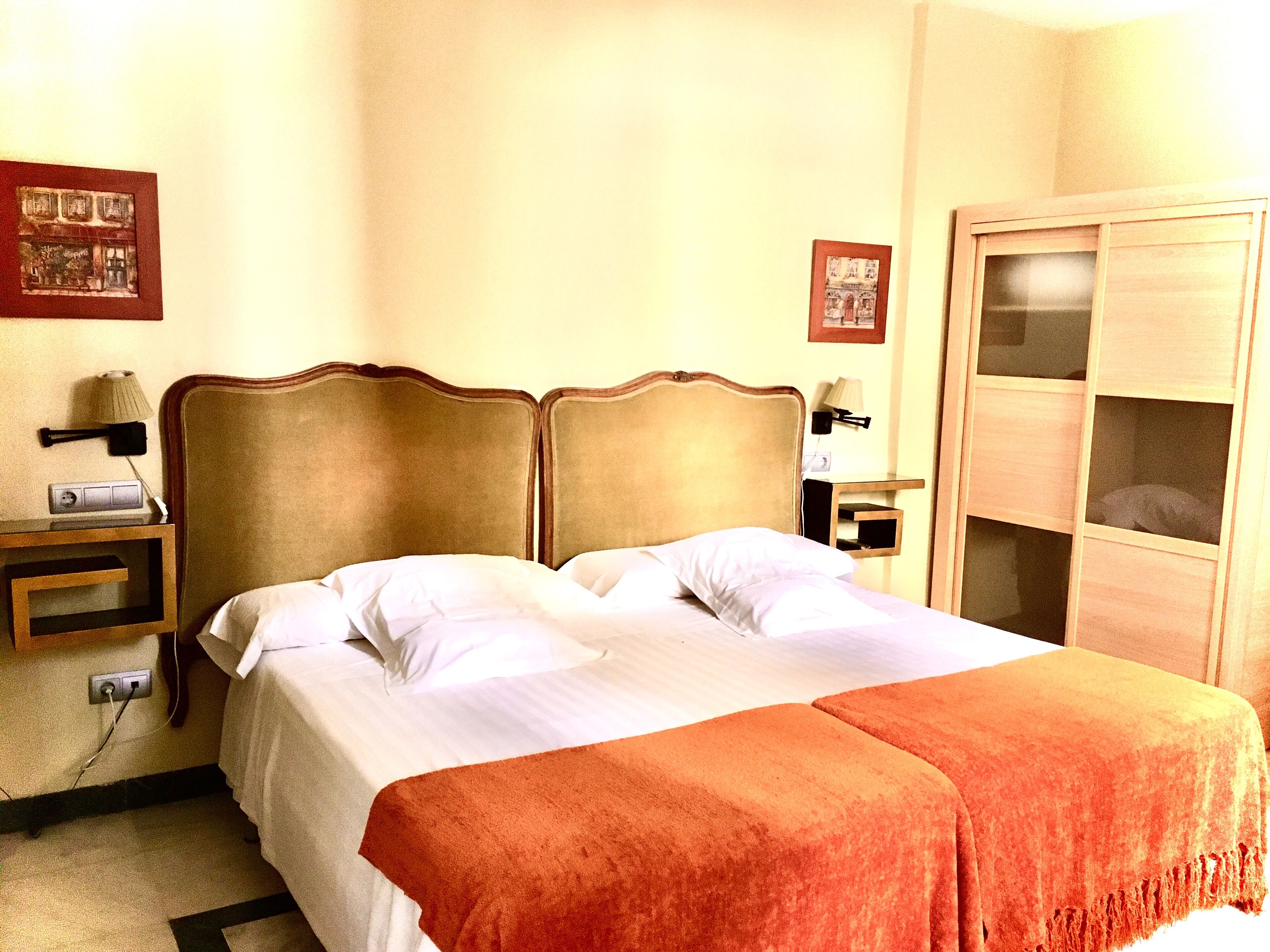A double bed with a wooden headboard, white linens, and an orange throw blanket. There is a nightstand with a lamp and a phone, and a wooden wardrobe with a mirror.