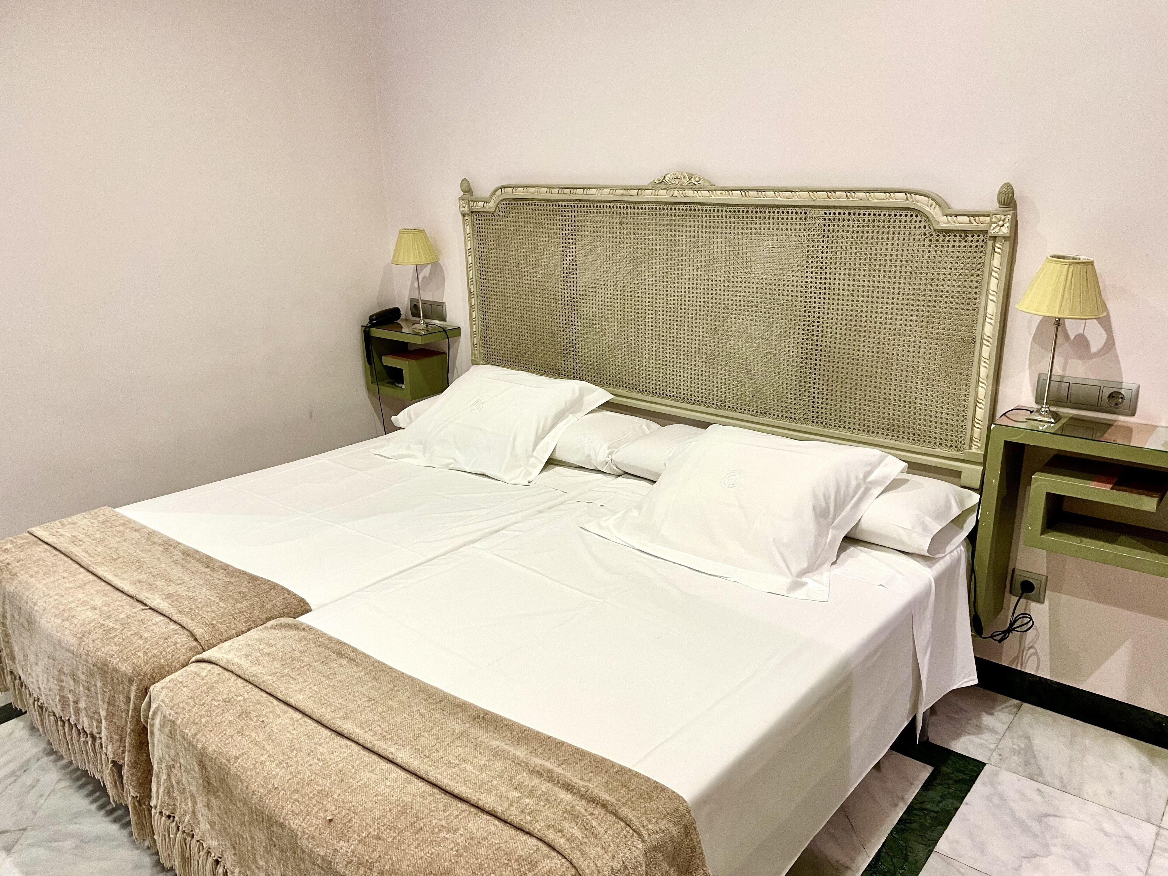 A double bed with white linens and a beige throw blanket, flanked by two bedside tables with lamps, in a room with a woven headboard and plain walls.