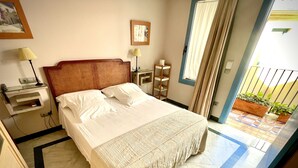 A neatly made bed with white linens, a wooden headboard, and a patterned bedspread. There is a small bedside table with a lamp, a framed picture on the wall, and a window with curtains. - Hotel Conde de Cárdenas (Córdoba)
