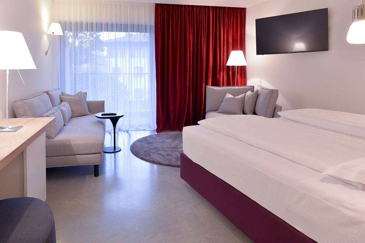 Habitación de hotel con cama, sofá, escritorio, televisor y ventana con cortinas.