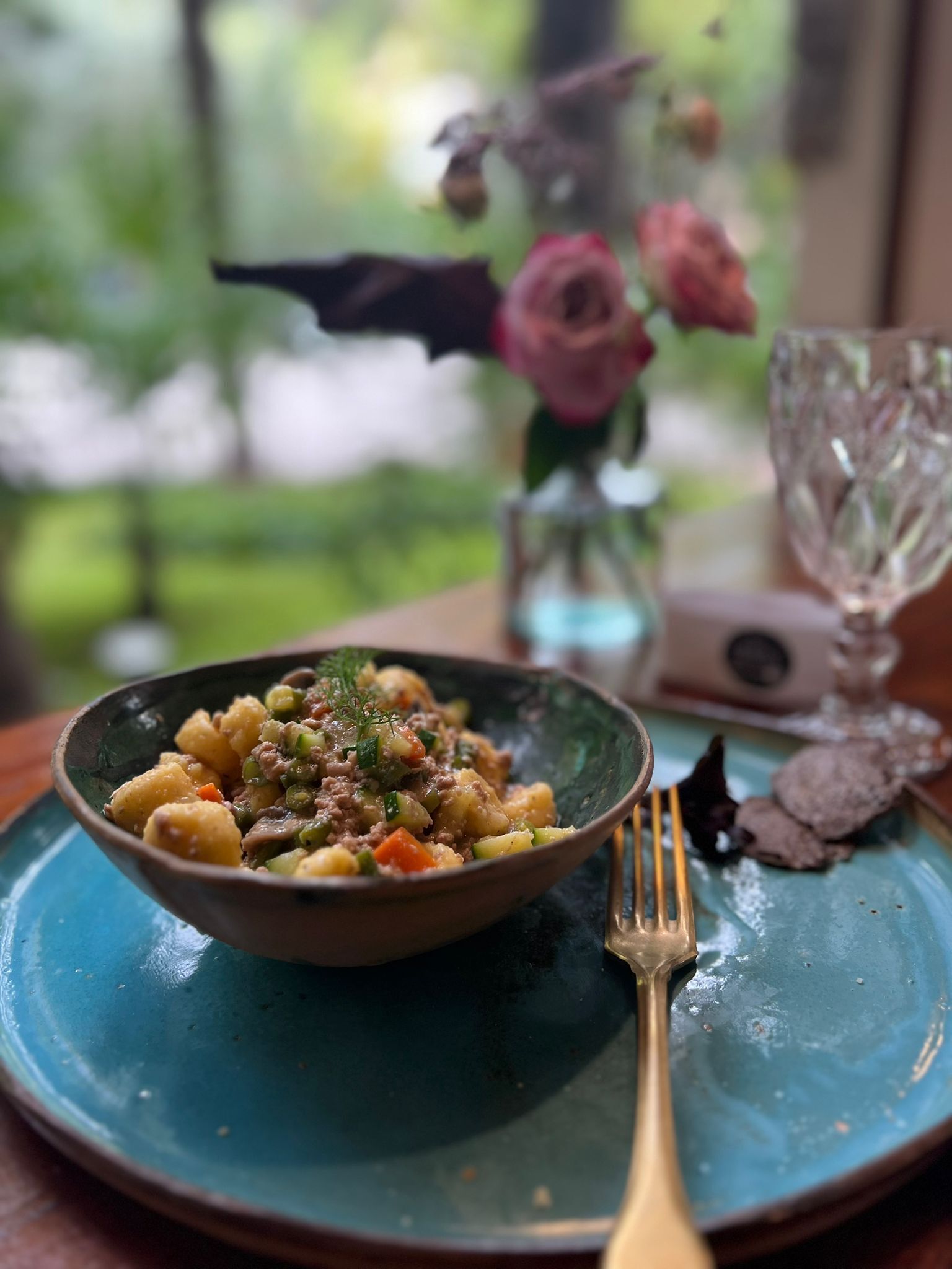 Un bol de nourriture sur une assiette bleue, avec une fourchette à côté, un verre et un vase avec des fleurs en arrière-plan.