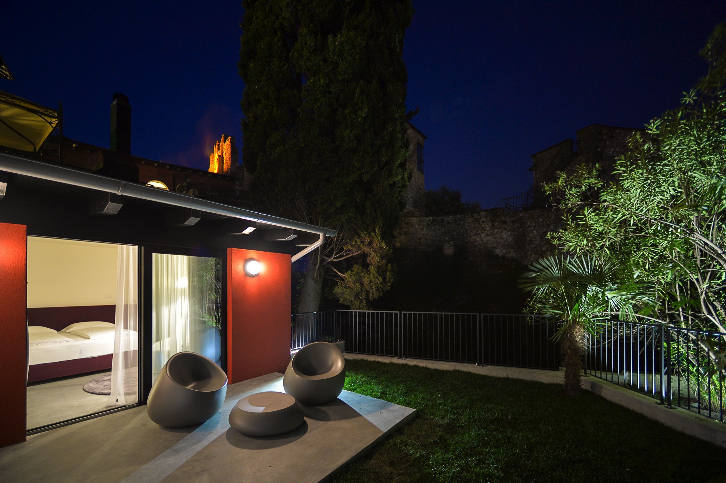 A modern house with a glass door entrance, a balcony with seating, and a well-lit garden area at night.