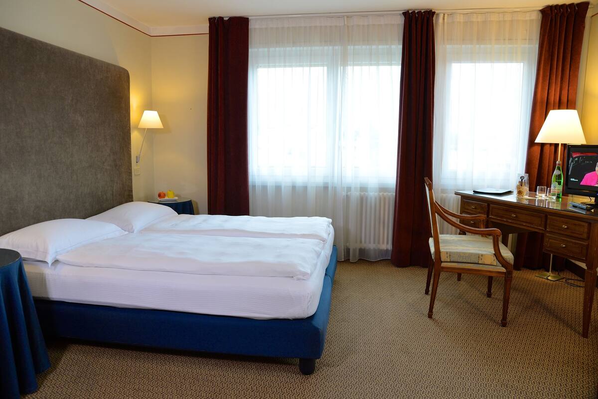 Habitación de hotel con cama doble, escritorio con silla, televisor y ventana con cortinas.