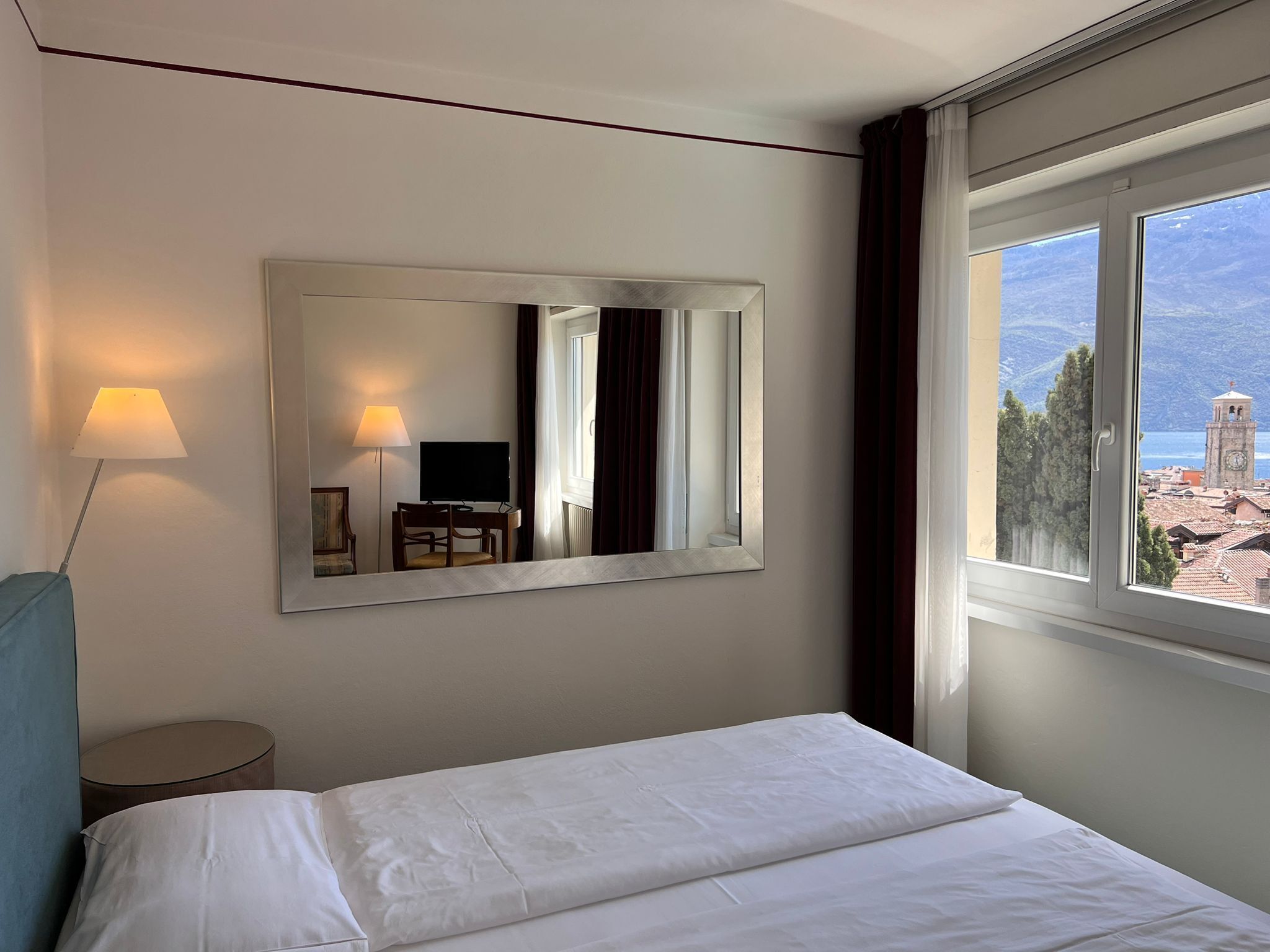 A hotel room with a bed, a desk with a chair, a TV, a lamp, and a large window with a view of a town and mountains.