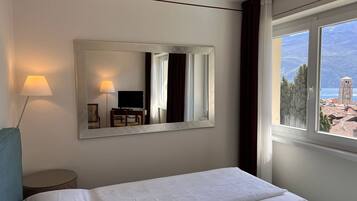 A hotel room with a bed, a desk with a chair, a TV, a lamp, and a large window with a view of a town and mountains.