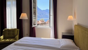 A hotel room with a bed, a chair, two lamps, and a window with a mountain and lake view.