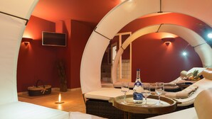 A modern, circular room with a bed, lounge chairs, a tray with glasses and a bottle, and a TV on the wall.