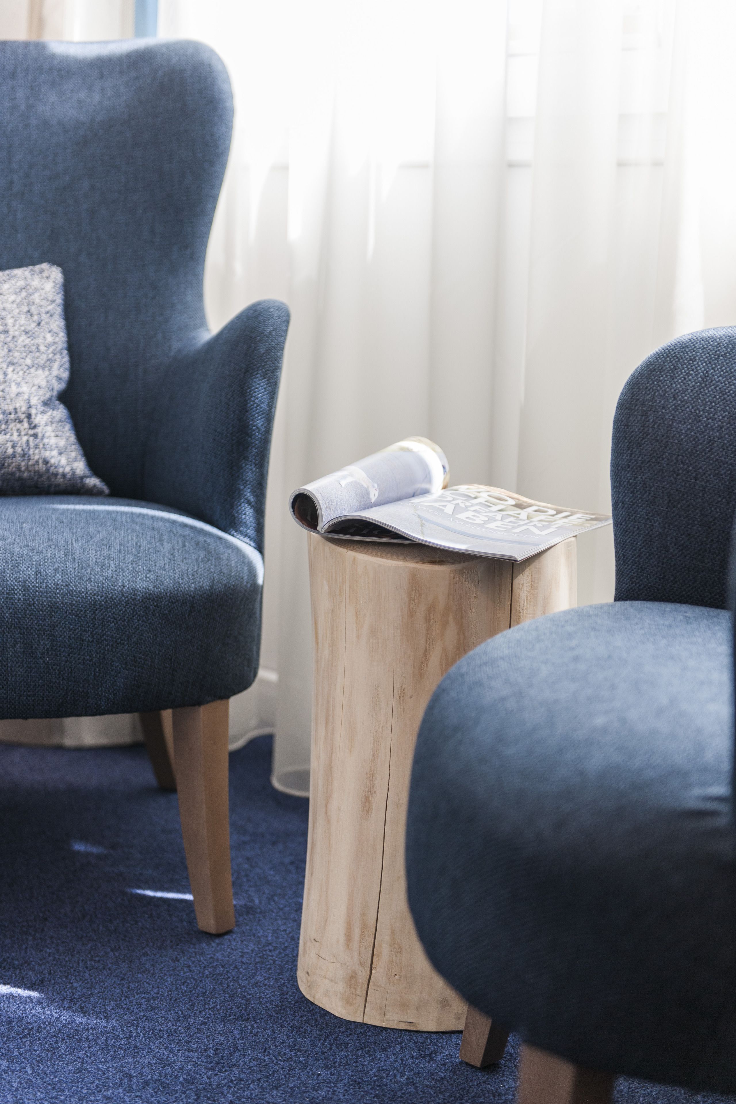 A room with a blue armchair, a wooden side table with a magazine, and a window with sheer curtains.