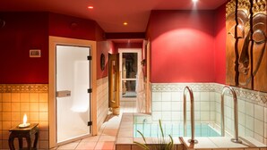 A hallway with a red wall, a wooden door, and a tiled floor leading to a spa area with a bathtub and a small table with a candle.