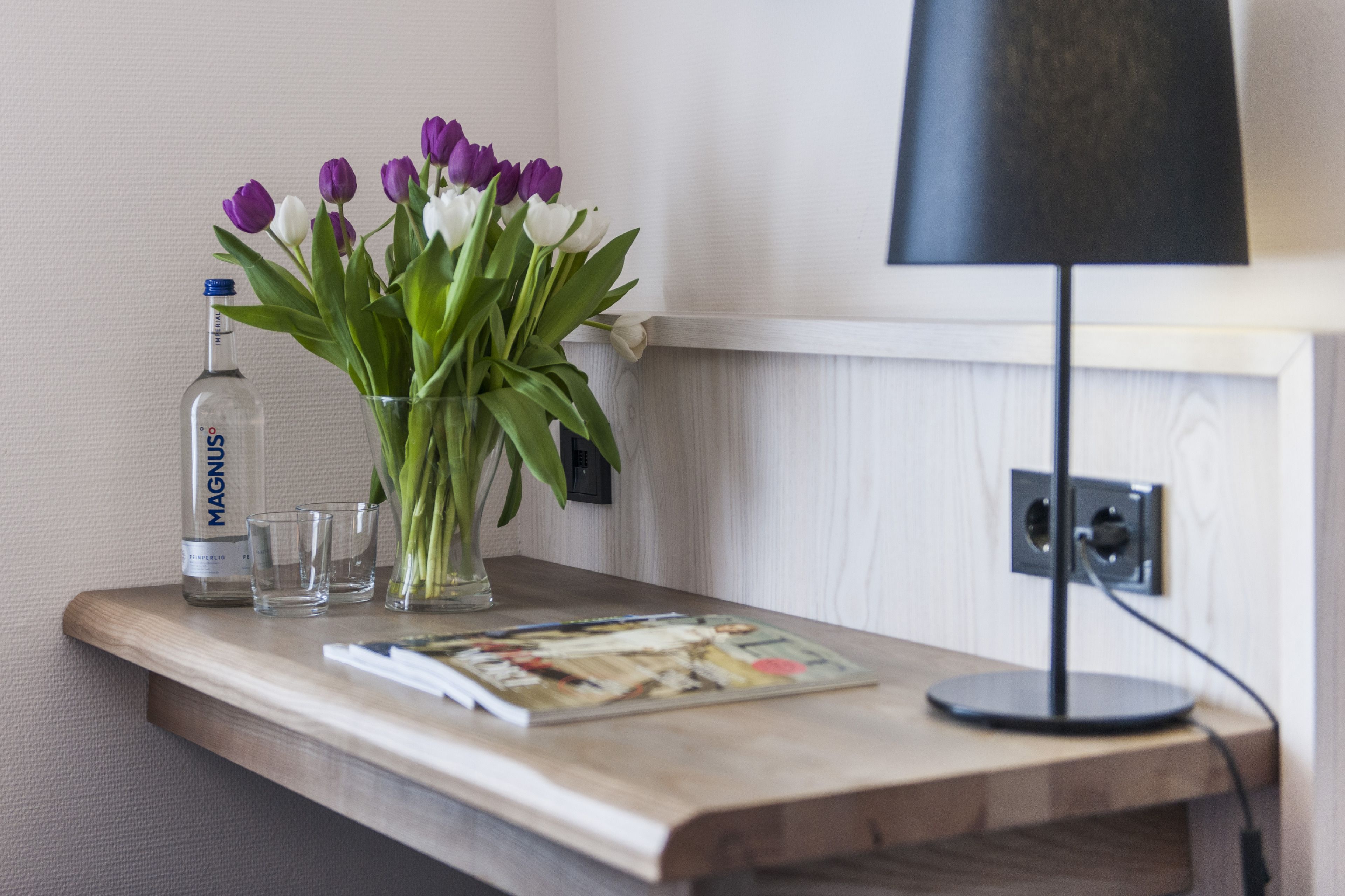 A wooden table with a magazine, a bottle of Magnus, and a vase of tulips.