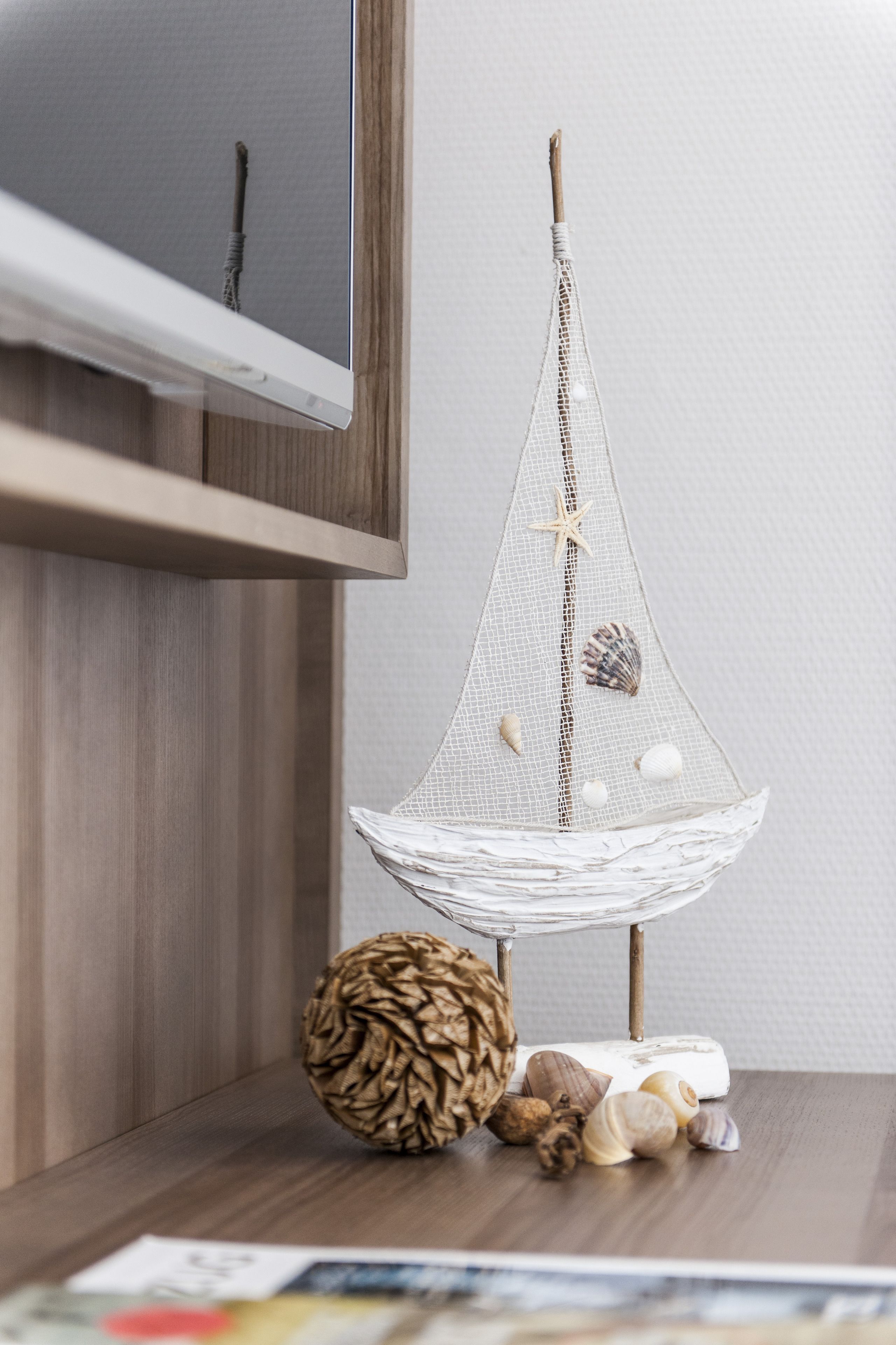 Uma pequena embarcação decorativa com conchas e uma estrela-do-mar, colocada sobre uma superfície de madeira ao lado de uma televisão.