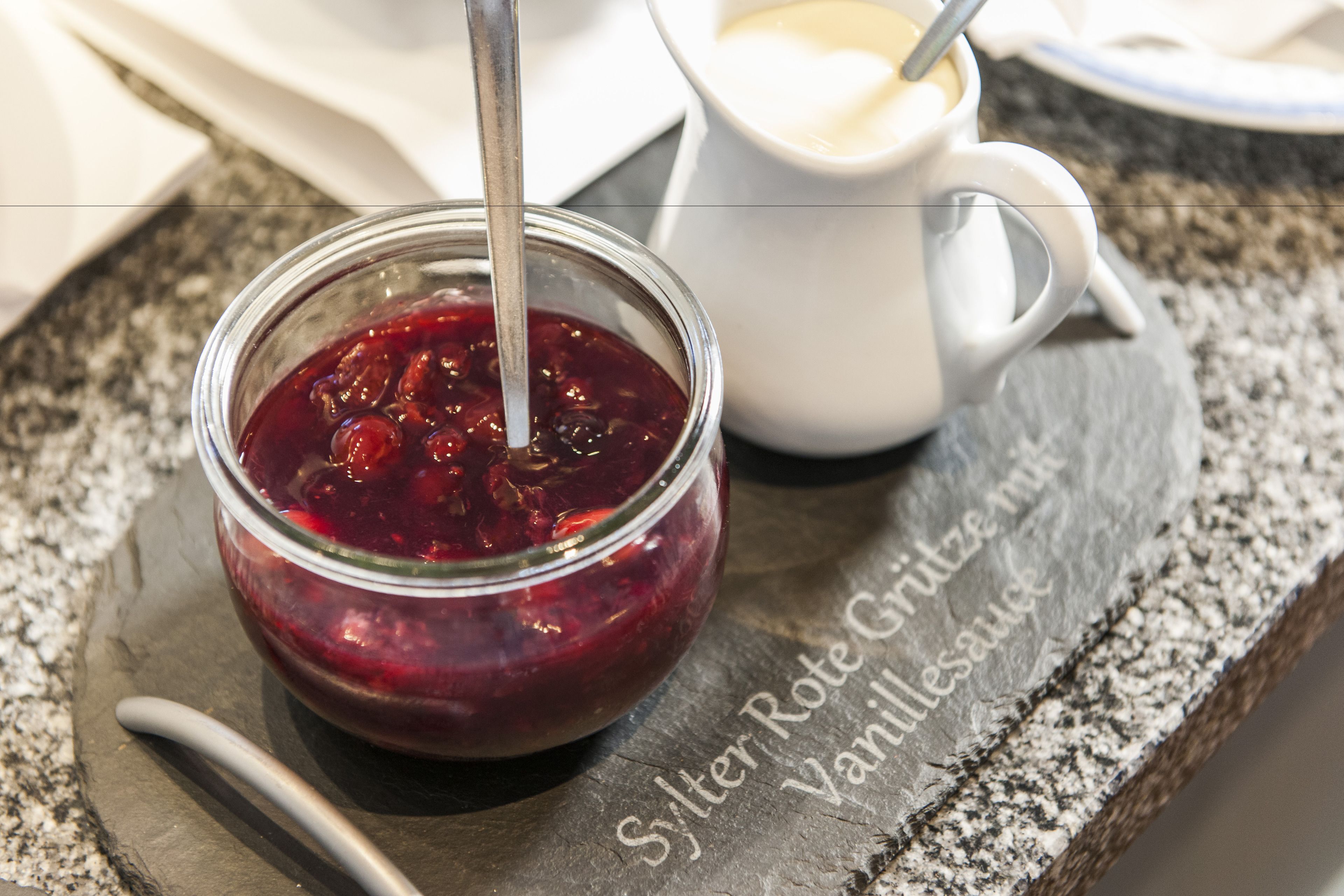 Et glas rødt marmelade med en ske, placeret på en skifer serveringsbakke med tekst.