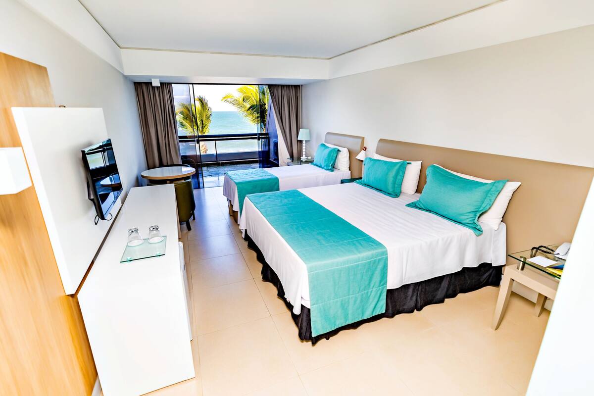 A hotel room with two beds, a desk, and a balcony with a view of the ocean.