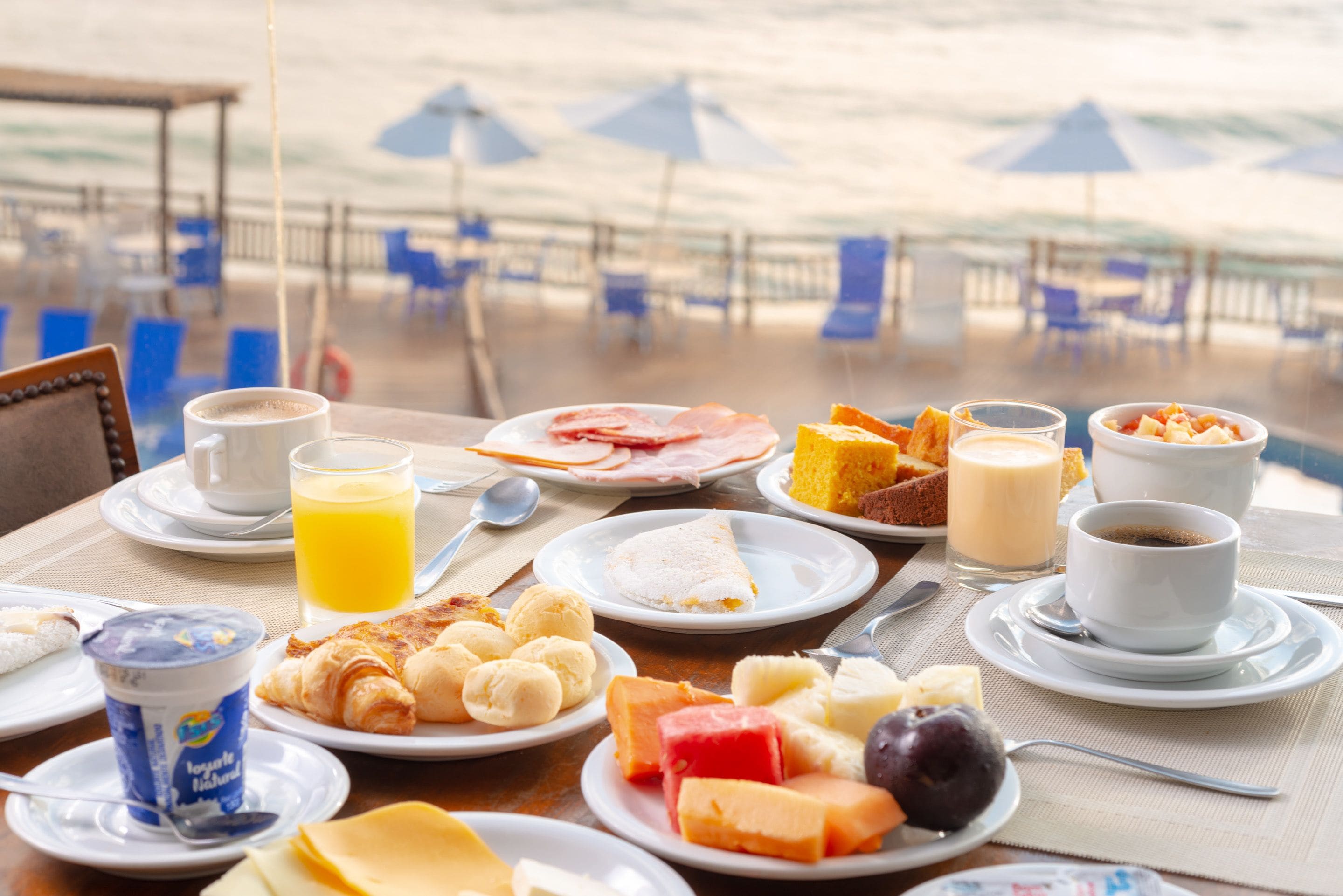 A breakfast table with various food items and drinks, overlooking a beach with umbrellas and chairs.