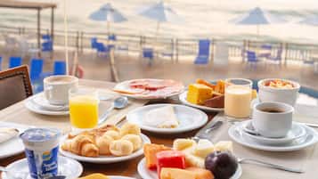 A breakfast table with various food items and drinks, overlooking a beach with umbrellas and chairs.